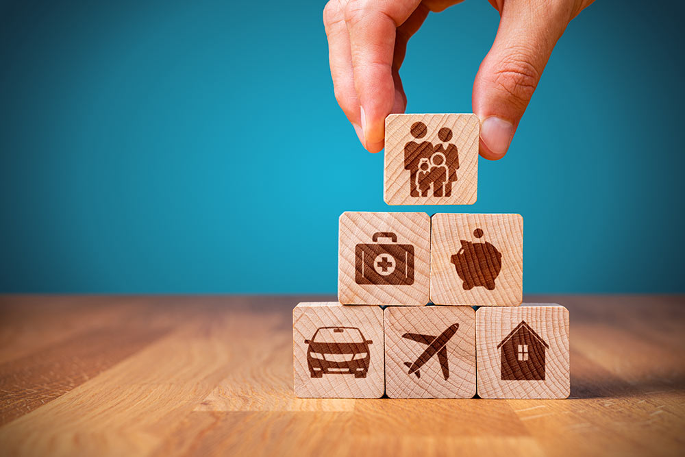 A person placing building blocks with a family, piggy bank, medicine bag, car, plane, and house